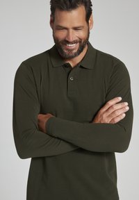 Langærmet polo shirt i mørkegrøn bomuldsstof. Har en klassisk krave, knaplukning og subtil tekstur.