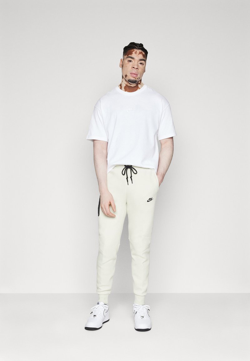 Weißes T-Shirt mit kurzen Ärmeln und kleinem Logo, kombiniert mit cremefarbenen Jogginghosen mit schwarzen Akzenten und einem Kordelzug in der Taille. Dazu werden weiße Sneakers getragen.