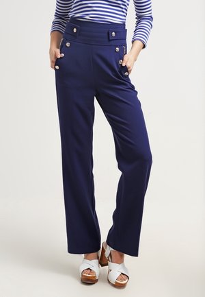 Femme portant un pantalon large taille haute bleu marine avec des boutons décoratifs et des sandales blanches à plateforme, mains dans les poches.