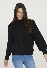 Pull en maille noir avec un col montant, présentant des motifs en relief et des accents de boutons dorés sur l'épaule. Coupe ample avec un ourlet côtelé.