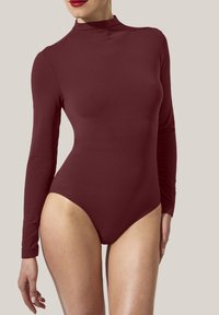 Kvinna iförd en figursydd, långärmad, höghalset bordeauxfärgad bodysuit som står mot en neutral ljus bakgrund.