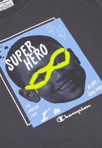 Camiseta gris con un estampado gráfico de un niño sonriente con gafas amarillas sobre un fondo azul, bordeado por líneas blancas y texto.