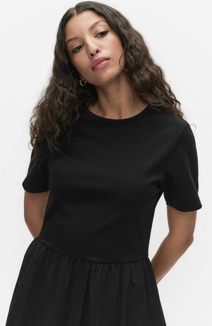 Kobieta o falowanych włosach, nosząca złote kolczyki koła i czarną sukienkę z krótkim rękawem, patrząca w dół na jednolitym tle.