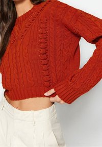 Pull court en maille torsadée couleur rouille avec ourlet et manches côtelés, porté avec un pantalon crème taille haute, main posée sur la hanche.