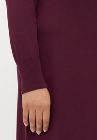Hand resting by side, wearing long-sleeve, ribbed cuff, dark purple knit garment, smooth fabric texture visible.