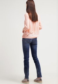 Blusa a maniche lunghe rosa chiaro abbinata a jeans skinny blu scuro e sneakers beige con suola testurizzata, vista da dietro.