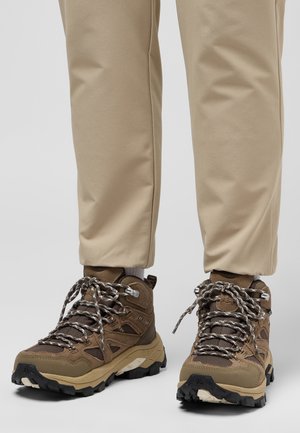 Bottes de randonnée marron avec semelles beige et crampons noirs portées avec un pantalon beige clair sur un fond gris.