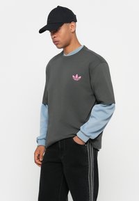 Uomo con cappellino nero, t-shirt grigio scuro con logo Adidas rosa sopra una maglietta a maniche lunghe blu chiaro, e pantaloni neri con strisce laterali, in piedi con una mano in tasca.