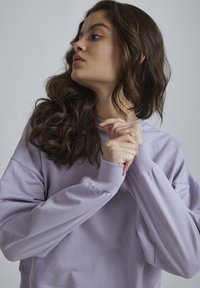 Felpa viola chiaro con polsini a coste e colletto rotondo, caratterizzata da una vestibilità comoda e una texture morbida e liscia. Il modello mostra il profilo di lato.