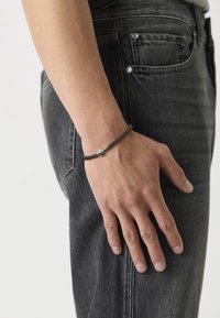 Bracelet noir tressé avec un accent en croix argentée, porté au poignet, complété par un jean en denim sombre. Design simple et minimaliste.