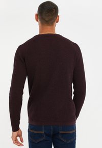 Maglione lavorato a maglia color bordeaux con collo rotondo e maniche lunghe. Presenta un motivo testurizzato, polsini e orlo a coste. Indossato con jeans blu.