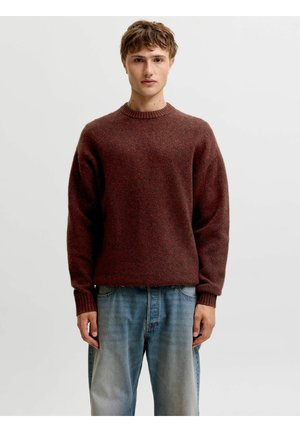 Jeune homme aux cheveux bruns courts portant un pull bordeaux et un jean bleu clair, debout devant un fond clair uni.