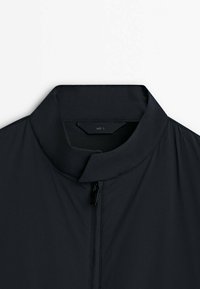 Veste noire avec un col montant et une fermeture éclair à l'avant, étiquette de taille grande visible à l'intérieur du col.