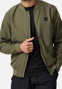 Homme portant une veste bomber vert olive par-dessus une chemise noire, tenant la fermeture éclair de la veste d'une main, pantalon noir visible en dessous.