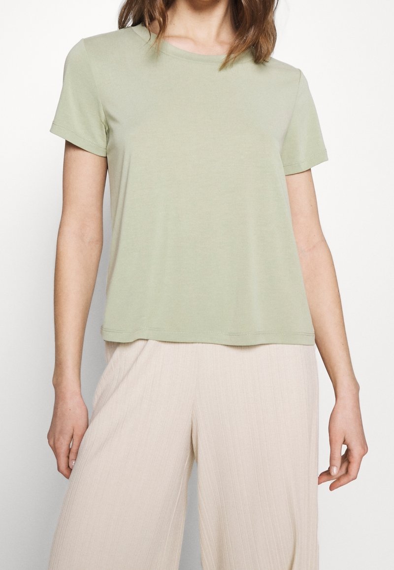 Femme portant un t-shirt vert clair à manches courtes et un pantalon beige à jambes larges, debout devant un fond blanc.