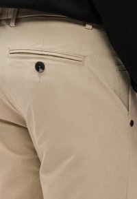 Gros plan sur un pantalon beige sur mesure montrant une poche arrière avec un seul bouton noir et le détail de la poche latérale.