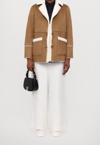 Hnědá umělá beránková bunda s bílým lemem, dvěma předními kapsami, hnědými knoflíky, nošena přes černou košili, v kombinaci s bílými kalhotami s širokými nohavicemi a černou taškou.