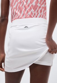 Άτομο που φοράει λευκή αθλητική φούστα και ροζ αμάνικη μπλούζα με σχέδια, ρυθμίζοντας το στρίφωμα της φούστας με το ένα χέρι.