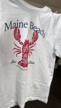 Balta T-kreklu ar sarkano omāru grafiku. Teksts augšpusē lasāms "Maine Beach", bet apakšpusē - "Labākais omārs Masačūsetsā."