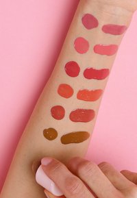 Farbproben verschiedener Lippenstiftfarben in kreisförmigen und ovalen Formen auf einem Arm mit Hautton, die Farbtöne von Pink bis Dunkelbraun auf einem pinkfarbenen Hintergrund zeigen.