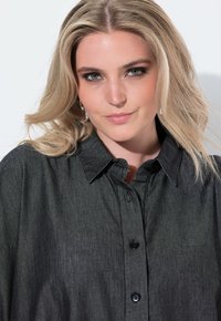Blonde Frau mit grünen Augen trägt ein dunkles, geknöpftes Hemd und hängende Ohrringe und lächelt leicht vor einem schlichten weißen Hintergrund.