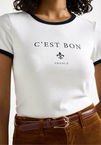 Witte katoenen t-shirt met marineblauwe afwerking, met de tekst "C'EST BON" en een fleur-de-lis afbeelding. Gecombineerd met bruine corduroybroeken en een leren riem.