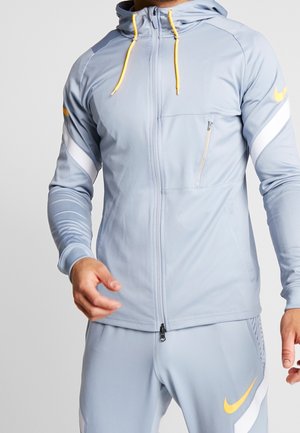 Homme portant un sweat à capuche zippé gris clair et un pantalon assorti avec des logos Nike jaunes et blancs et des cordons jaunes sur la capuche.