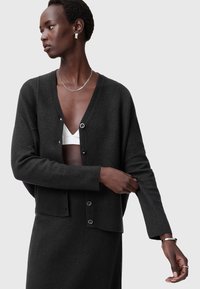 Mörkgrå kofta i stickad kvalitet, med V-ringning, knappar fram och sänkt axel. Bärs över en vit topp.