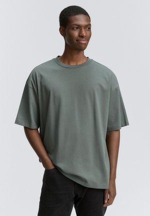 Camiseta de manga corta, holgada, en tela de algodón verde apagado. Presenta un cuello redondo y un sutil brillo. Combinada con pantalones oscuros.