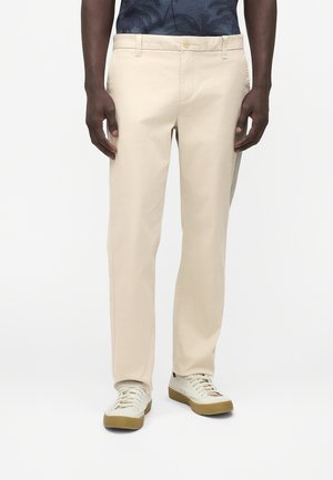 Mann trägt beige gerade geschnittene Hose, weiße Sneakers mit Gummisohle und ein dunkelblau gemustertes Hemd, stehend vor einem einfarbigen Hintergrund.