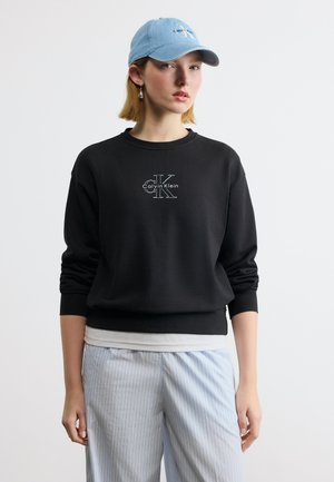 Junge Person trägt schwarzen Calvin Klein Pullover, hellblauen Cap und hellgraue gestreifte Hose und steht vor einem einfarbigen Hintergrund.