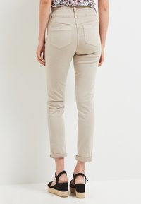 Femme portant un pantalon beige à ourlets et des sandales à semelle compensée noires, vue de dos contre un fond blanc uni.
