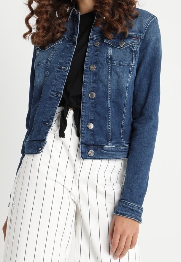 Tummansininen farkkutakki, jossa on klassinen kaulus, nappikiinnitys ja kaksi rintataskua. Takki on päällä mustan paidan ja raidallisten valkoisten housujen kanssa.