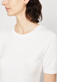 T-shirt alb din bumbac cu guler rotund și mâneci scurte, având o textură netedă și un design minimalist, purtat cu cercei rotunzi din aur.