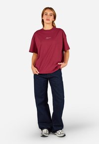 Burgundiju kreklu ar īsām piedurknēm un izšūtu logotipu, kombinējot ar tumši zilu platās kājas biksēm un pelēkiem sporta apaviem.