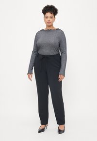 Siva teksturirana majica dugih rukava sa oštrim izrezom, u kombinaciji sa crnim pantalonicama visokog struka sa vezicom, upotpunjeno crnim cipelama na visoku petu.
