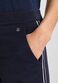 Mörkblå texturerade byxor med en svart metall-logotyp på sidofickan. Har en vit och röd rand längs kanten av fickan.