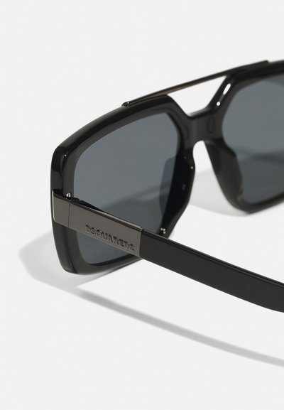 Mustad mustriga prillid, millel on massiivne ruudukujuline raam, tumedad läätsed ja metallist detailid tempel, kus on brändinimi "DSQUARED2."