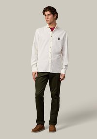 Camisa blanca de botones con mangas largas, que presenta un pequeño emblema en el pecho. Combinada con pantalones verde oscuro y calzado marrón.
