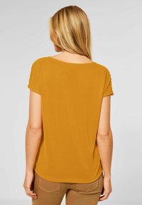 Kvinna med långt blont hår som bär en senapsgul kortärmad topp och bruna byxor, visad bakifrån mot en enhetlig bakgrund.