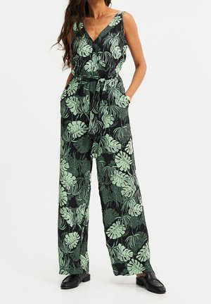Sort jumpsuit med grønt løvmønster, V-hals, vide ben, bånd i taljen og sidelommer. Glat stof og afslappet pasform. Sorte sko.