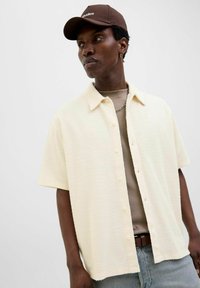 Camisa de botones de manga corta, texturada en crema, con cuello extendido, sobre una blusa taupe, combinada con jeans azul claro. Se accessoriza con una gorra marrón.