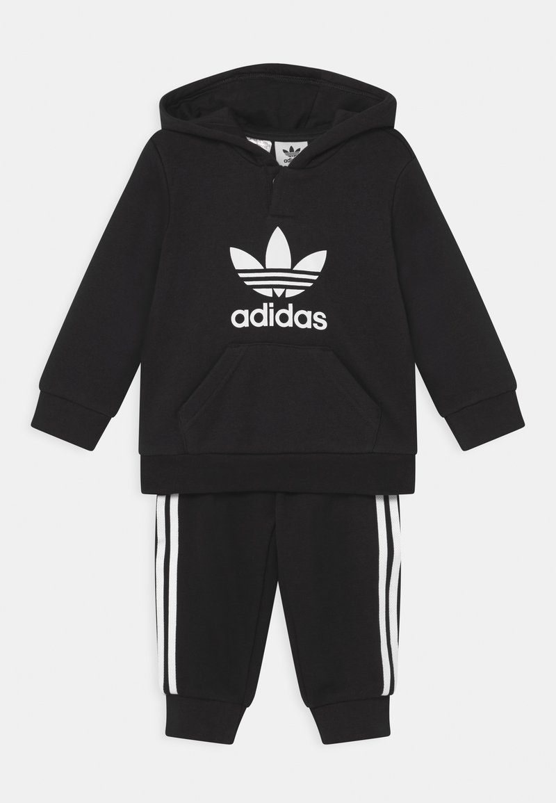 Čierna mikina Adidas s bielym logom a predným vreckom, skombinovaná s čiernymi teplákmi s bielymi pruhmi po bokoch.