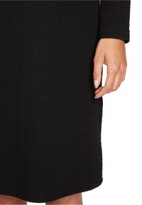 Svart klänning med ribbad textur, långa ärmar och knälång nederkant. En hand är placerad nära sidan av sömmen.