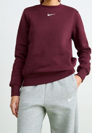Person iført en mørkerød sweatshirt og lysegrå joggingbukser, begge med hvide Nike swoosh-logoer, hånd i lommen.
