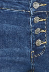 Jean en denim bleu à taille haute, avec quatre boutons argentés et une couture jaune contrastante le long des coutures.