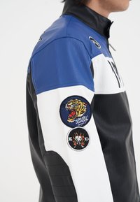 Herrlädermotorcykeljacka med blå, svart och vita paneler; tre broderade märken; och en hög krage.