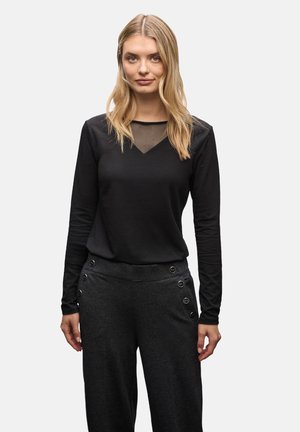 Blonde vrouw in een zwart lange mouw top met een doorschijnende halslijn en donkergrijze broek met knoopdetails aan de zijkanten.