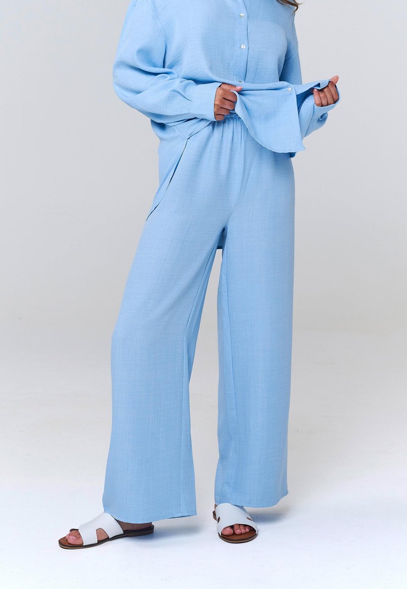 Femme portant un pantalon ample bleu clair avec un haut boutonné assorti, tenant l'ourlet du haut, et des sandales blanches ouvertes sur un fond neutre.