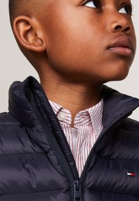 Giacca imbottita blu marina con zip frontale e colletto alto. Sotto, si vede una camicia bianca con strisce rosse e colletto button-down.